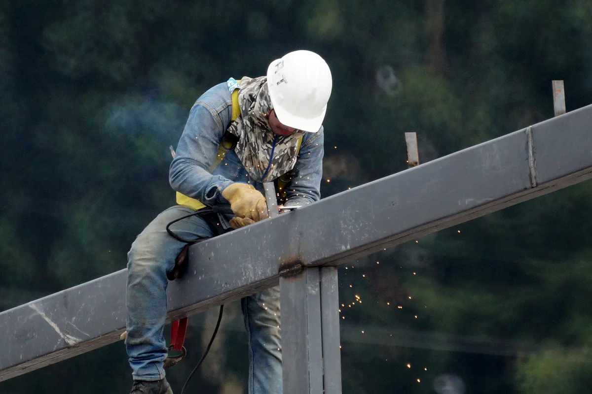 A construction worker in a white hard hat welding a steel beam overhead, sparks falling, neck craned upward.
