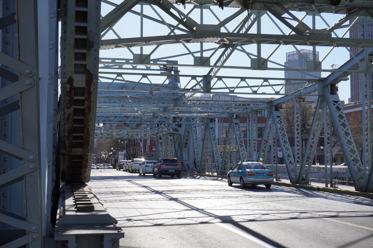 Cars crossing a steel truss bridge in daylight with a city skyline beyond.