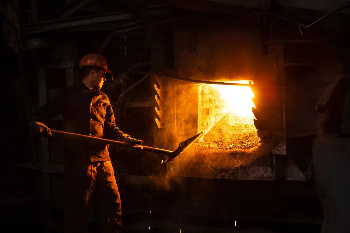 A steelworker in a hard hat tending a glowing industrial furnace.