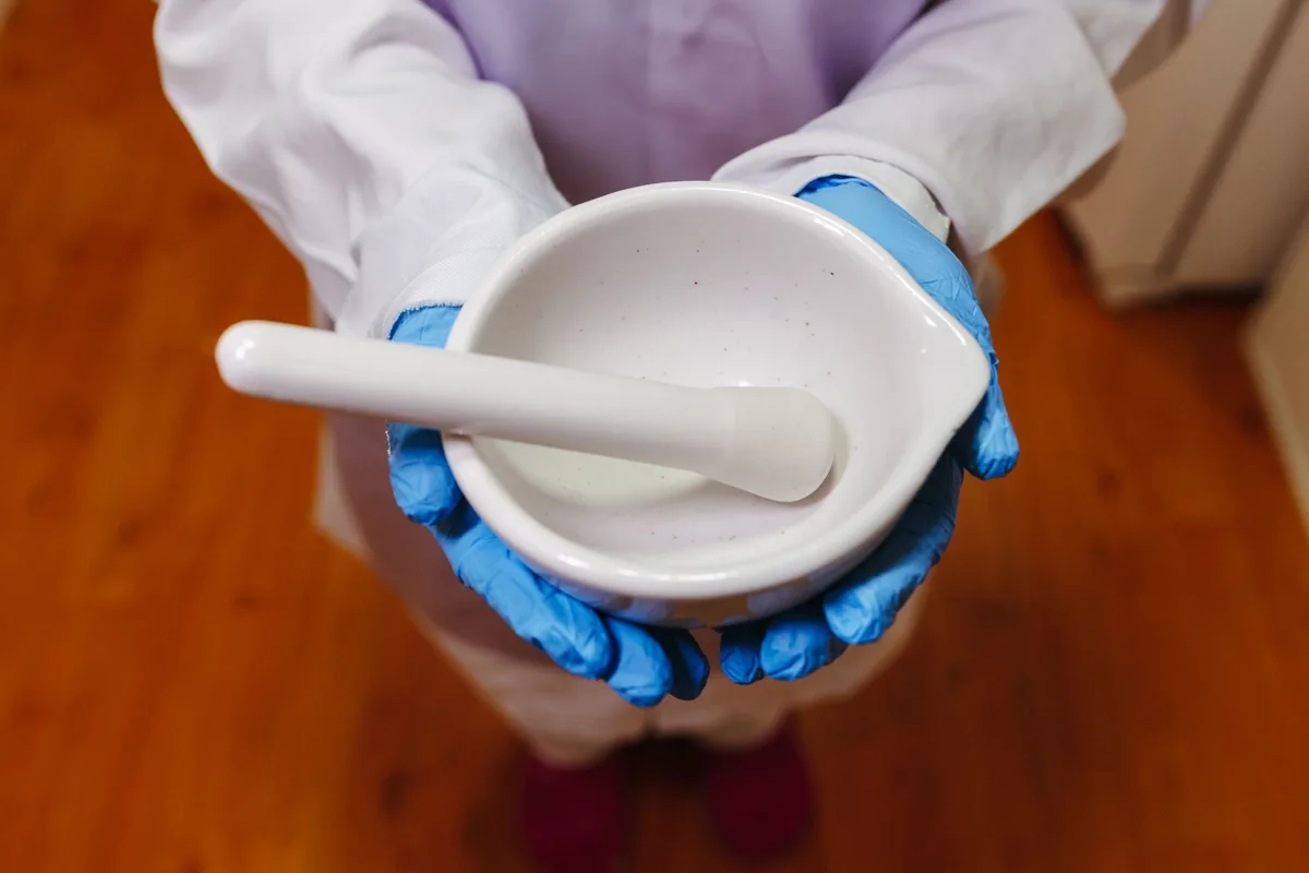A pharmacist in blue gloves holding a white mortar and pestle used for compounding.