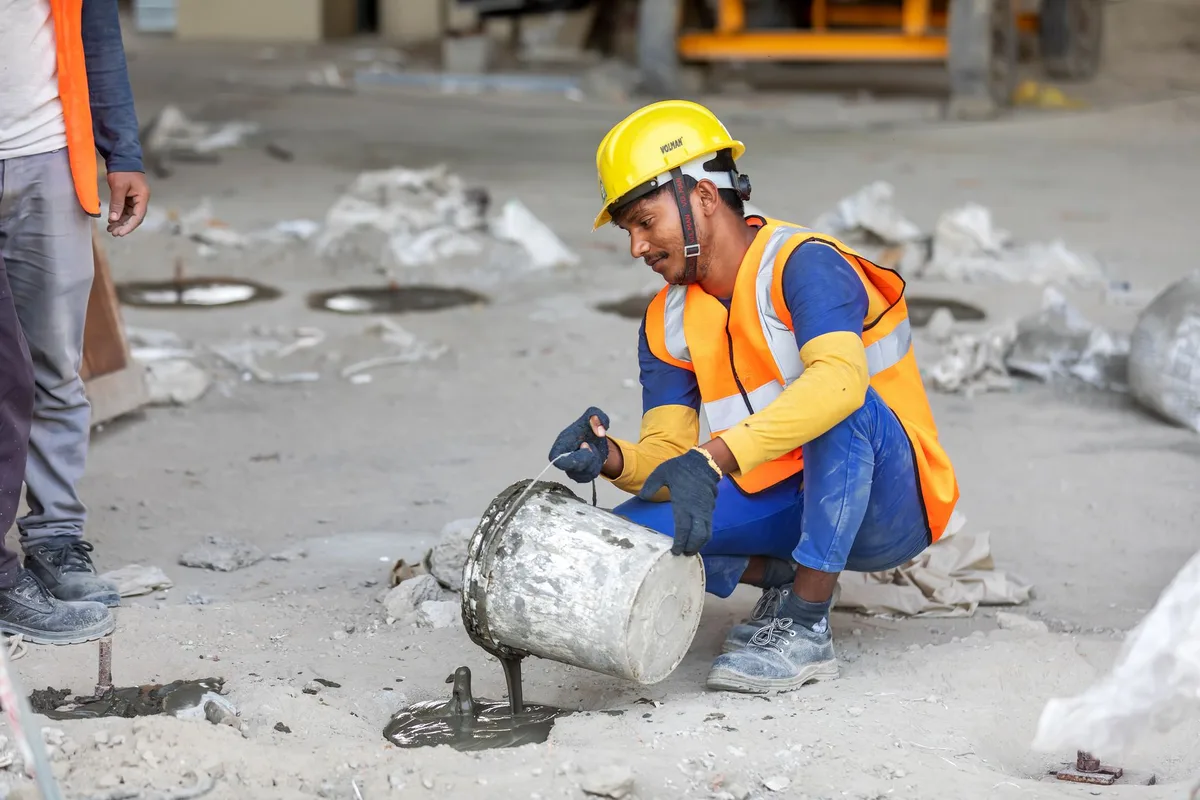 A construction worker in a hard hat and safety vest kneeling on concrete to pour cement.