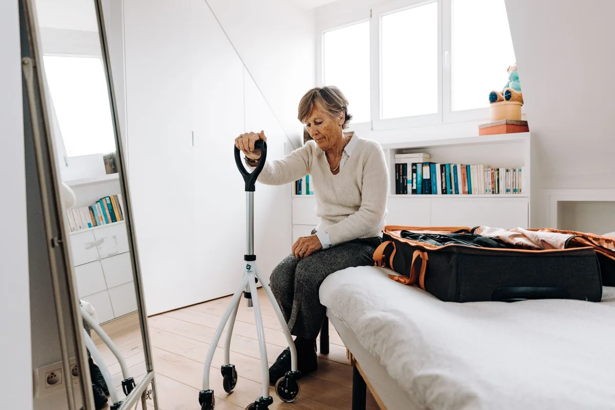 An older woman sitting on the edge of a bed with a rolling walker in front of her, a packed suitcase beside her.