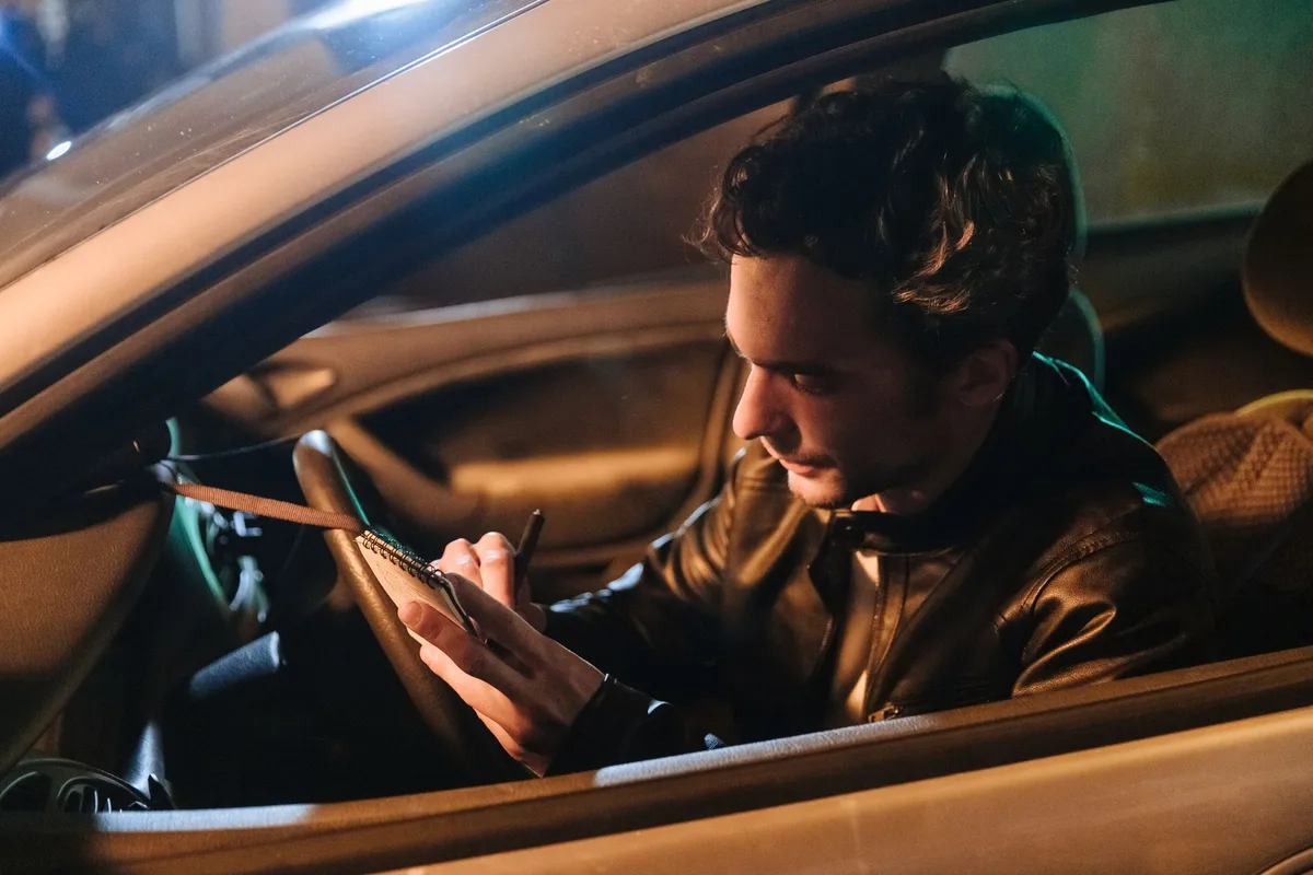 A man seated in a parked car at night writing notes in a small notebook.