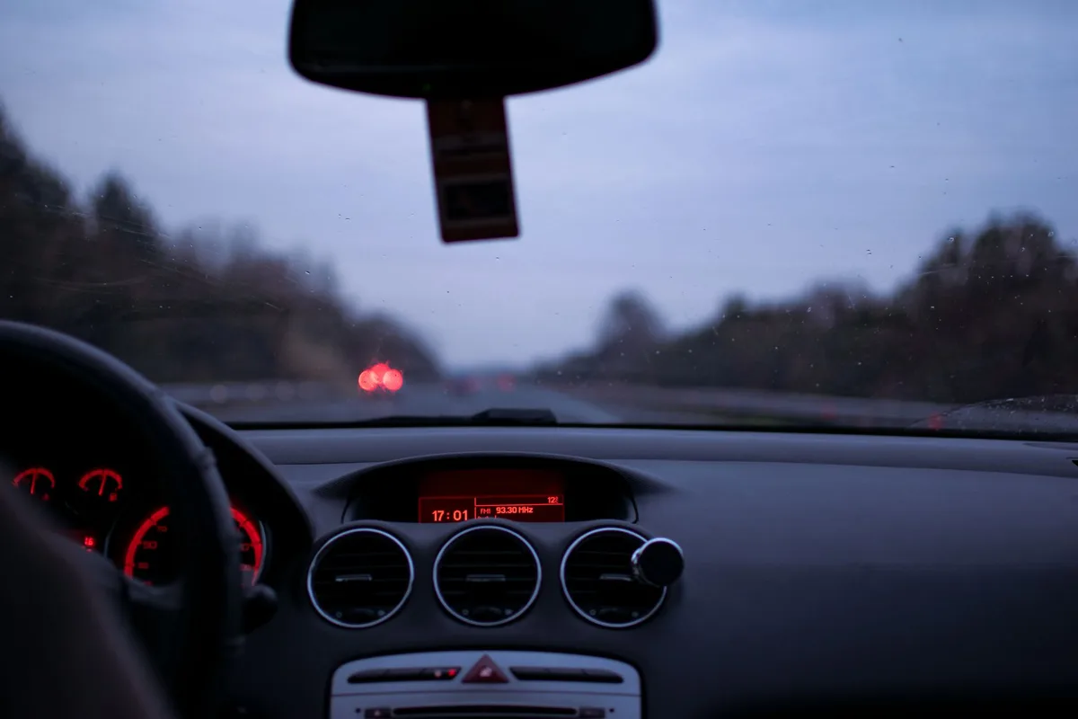 View over a car dashboard through the windshield of a highway at twilight, taillights ahead.
