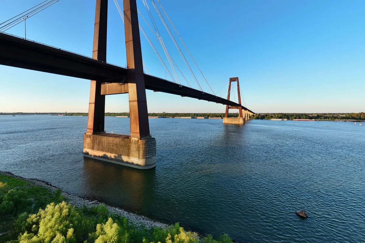 A cable-stayed bridge crossing the Mississippi River at sunset, seen from the riverbank.