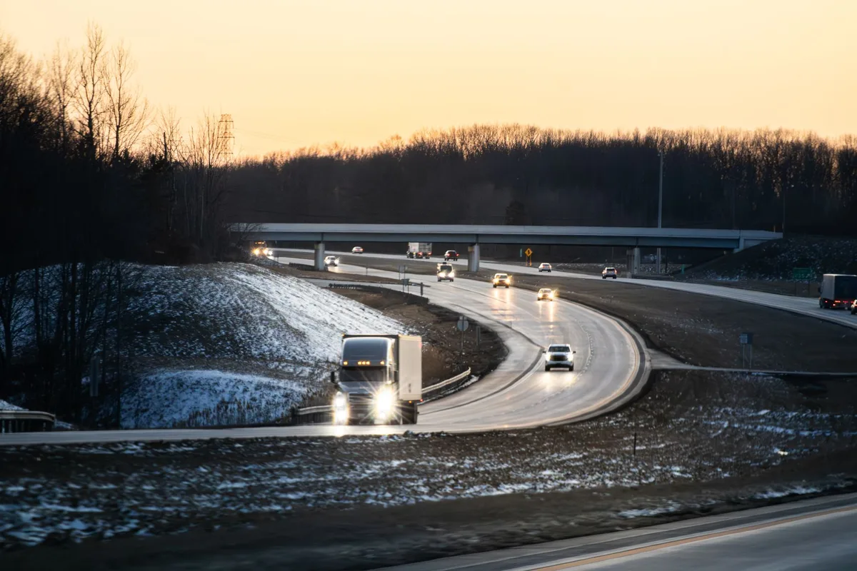 Cars and trucks on a multi-lane highway at dusk with light snow on the roadside.