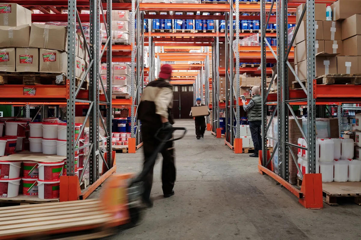 A worker pulling a loaded pallet jack down a distribution warehouse aisle.