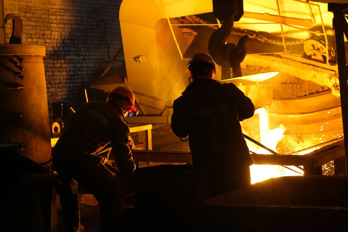 Two steelworkers in protective gear beside a furnace pouring molten metal.