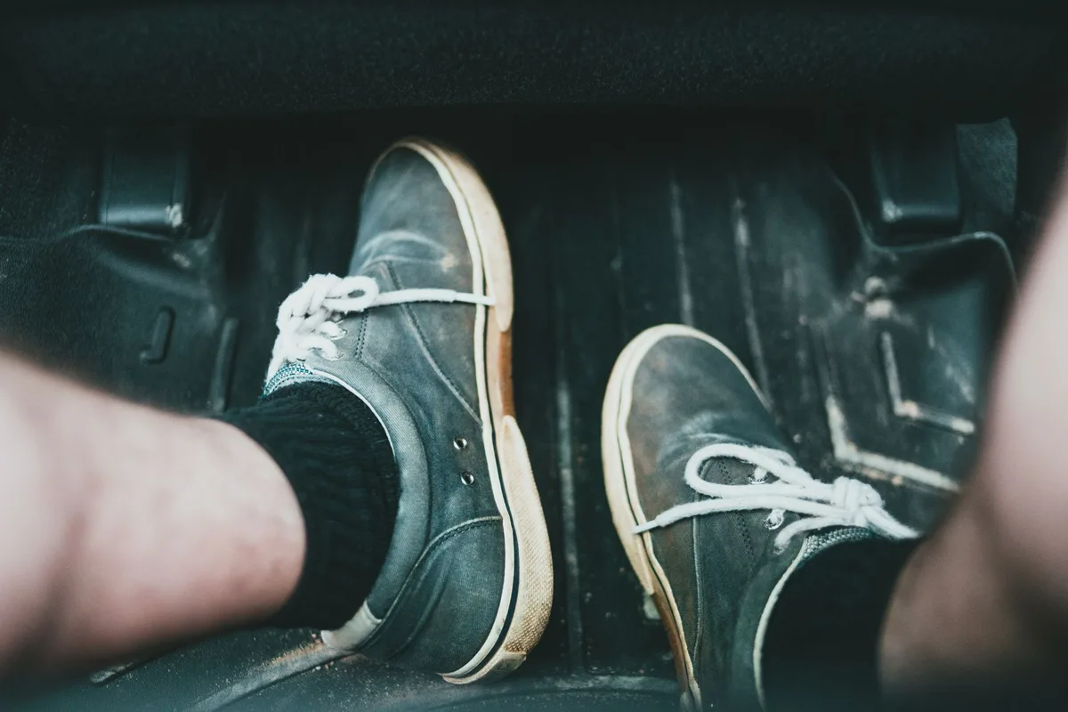 Close view of a driver's sneakers on a car floor mat by the pedals.