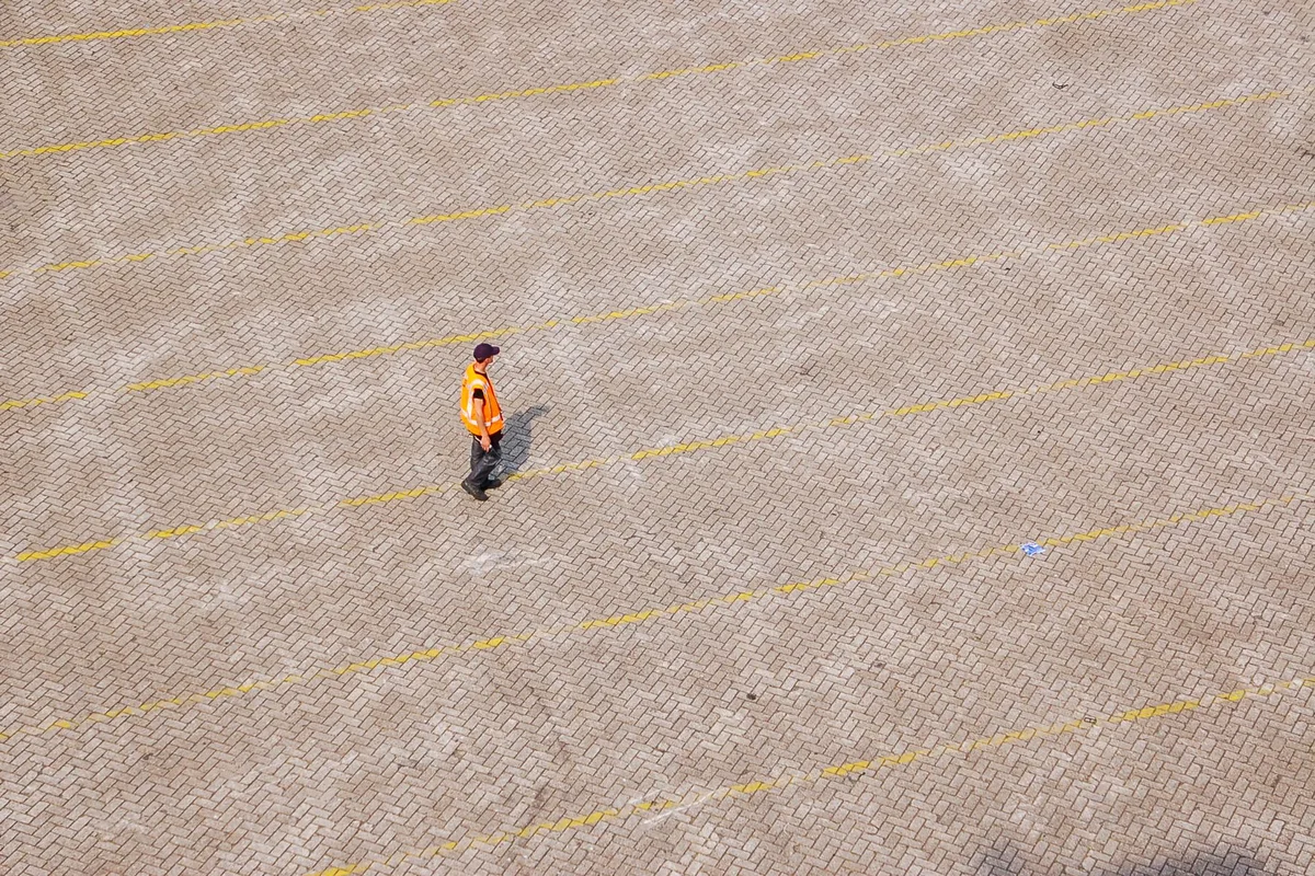 A lone worker in a high-visibility vest crossing an empty parking lot, seen from above.