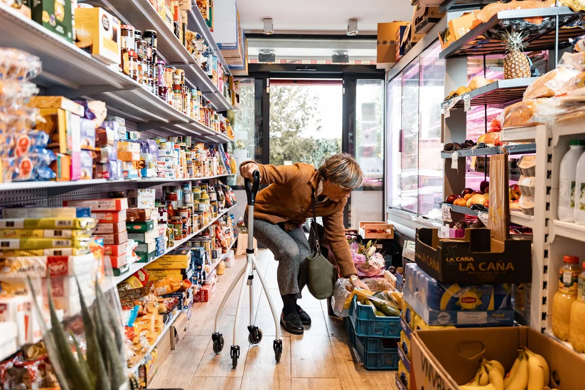 An older woman leaning on a rolling walker while choosing produce in a grocery aisle.