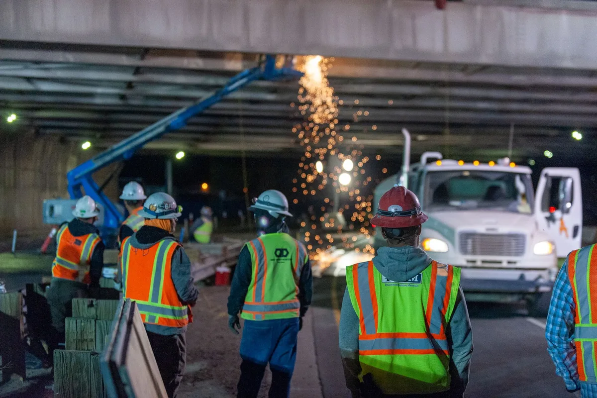 Road construction crew in hard hats and safety vests working at night under a bridge, sparks falling behind them.