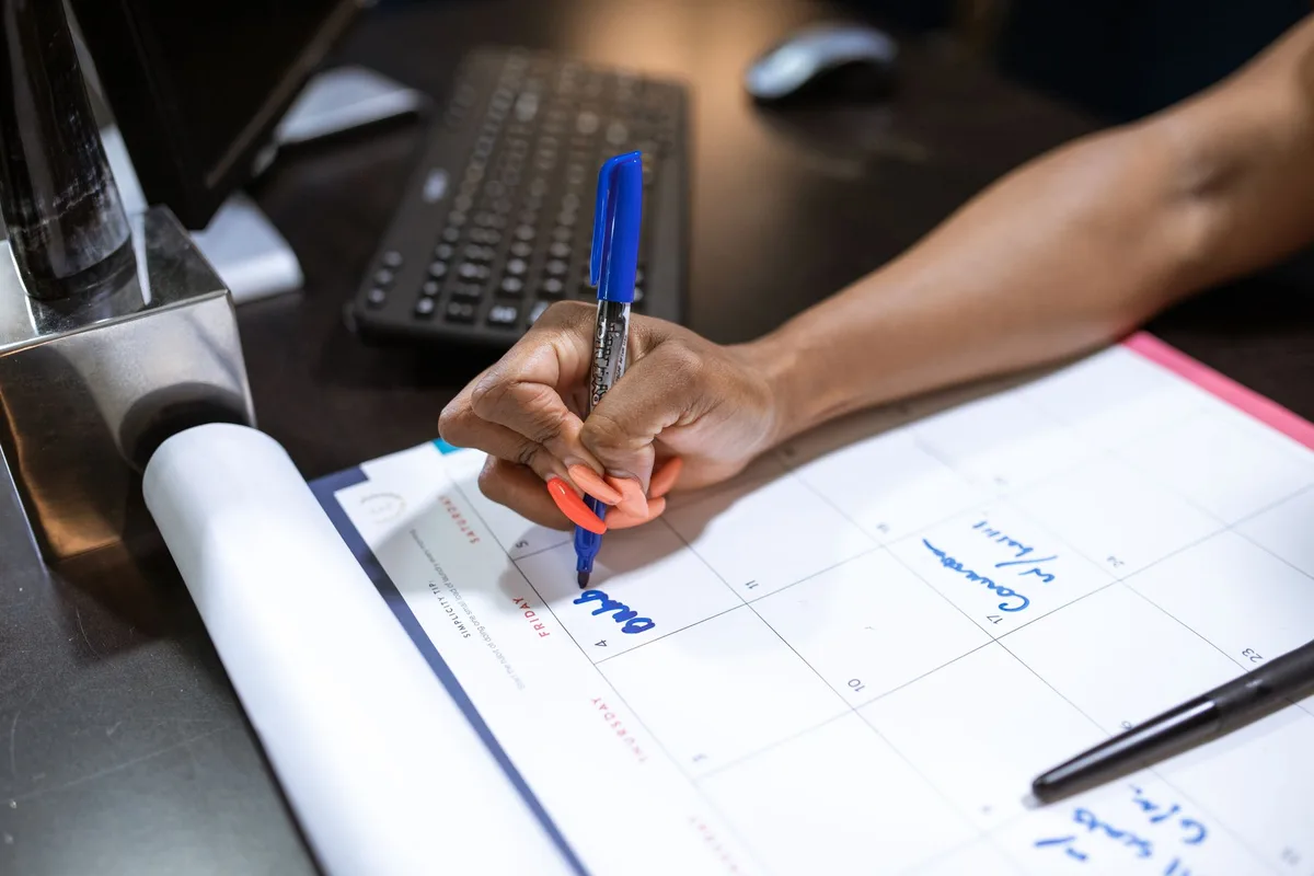 A hand writing appointment notes on a paper desk calendar with a blue marker.
