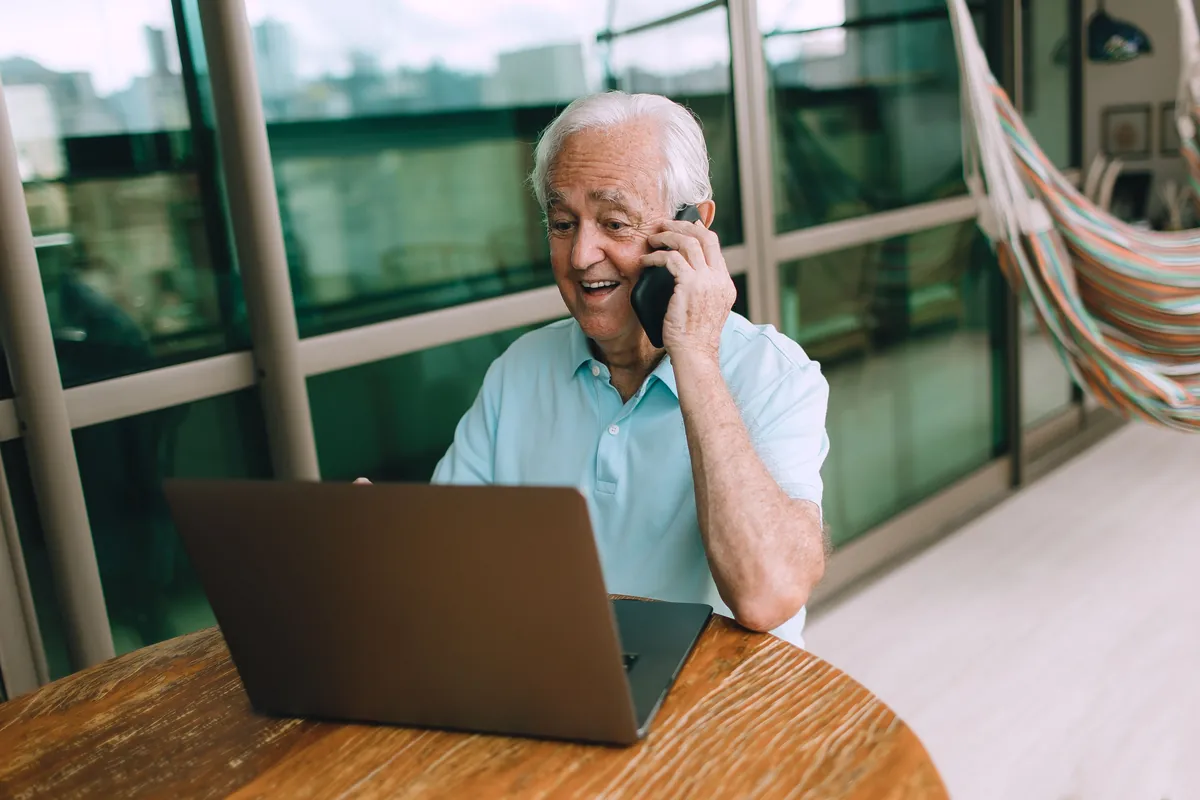 An older man talking on his phone while sitting at a laptop at home.