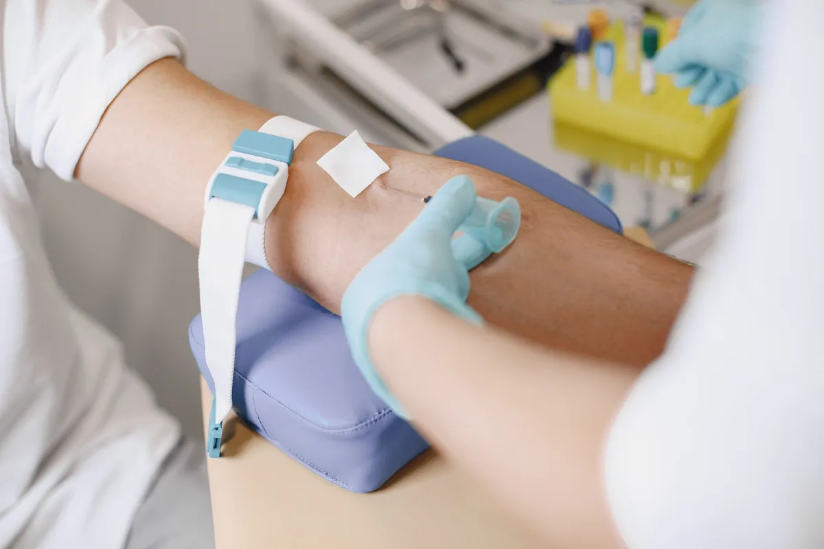 A gloved clinician drawing blood from a patient's arm, with collection tubes in a rack nearby.